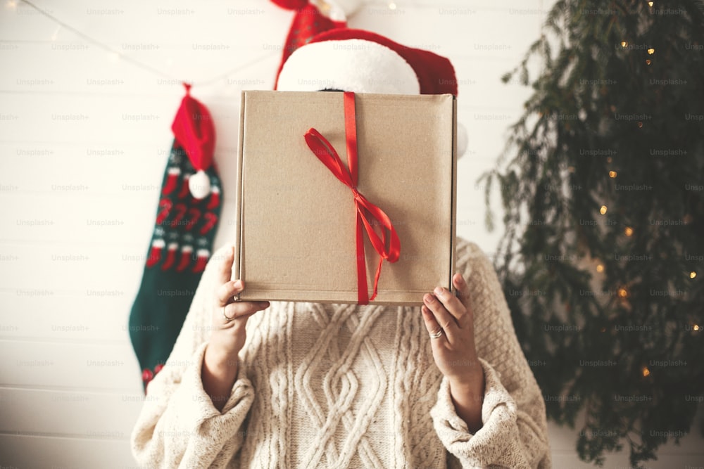 Menina feliz elegante no chapéu de Papai Noel segurando a caixa de presente de Natal na frente do rosto no fundo da árvore de Natal moderna, luzes e meias. Mulher hipster nova no suéter aconchegante segurando presente