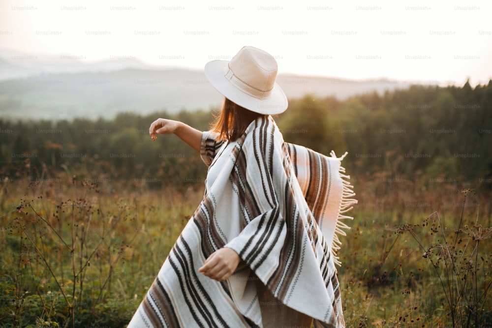 牧草地の雰囲気のある夕暮れ時に日当たりの良い光の中を歩くスタイリッシュな自由奔放に生きる女の子。ポンチョと帽子をかぶった幸せなヒップスターの女性が夜の山を旅しています。テキスト用のスペース