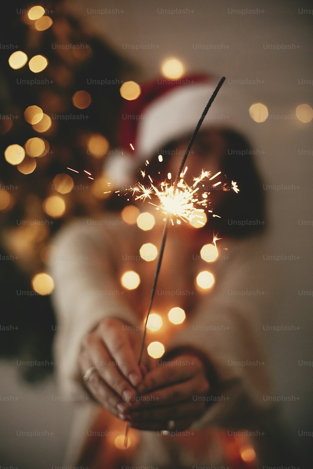 Happy New Year eve party atmosphere. Sparkler burning in hand of stylish girl in santa hat on background of modern christmas tree light in dark room. Woman with fireworks. Happy Holidays