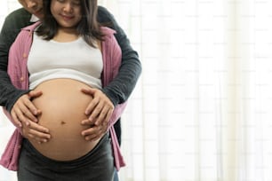 Pregnant couple of husband and wife feels love and relax at home. Young expecting woman holds baby in pregnant belly. Father take care of pregnant mother. Concept of maternity and pregnancy care.