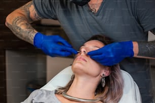 Portrait of a woman getting her nose pierced. Man showing a process of piercing nose with steril neadle and latex gloves. Nostril Piercing Procedure