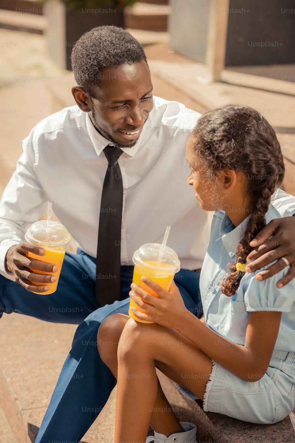 Friendly conversation. Emotional girl holding glass with orange juice and sitting near her daddy on stairs