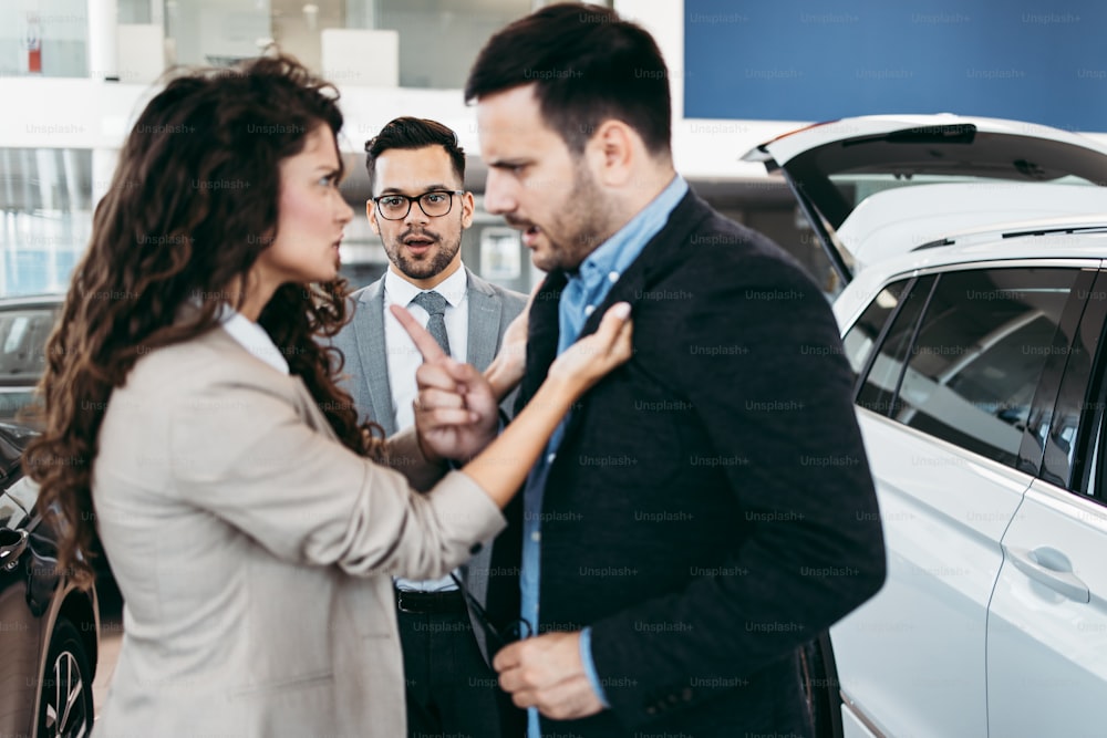Couple d’âge moyen se disputant lors de l’achat d’une voiture dans une salle d’exposition automobile. Le vendeur de voitures les aide à prendre la bonne décision.