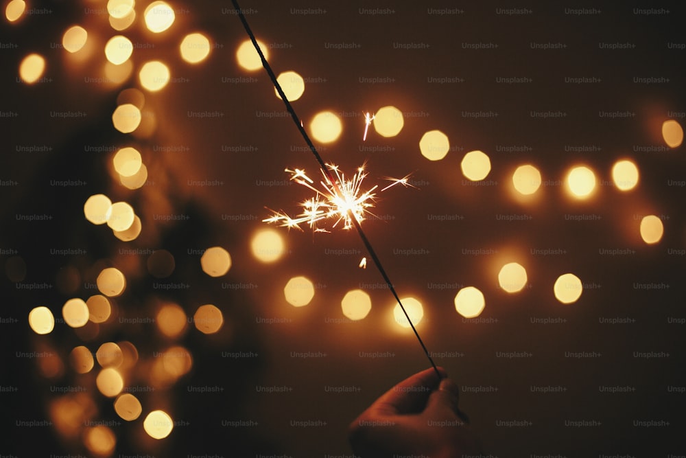 Glowing sparkler in hand on background of golden christmas tree lights, celebration in dark festive room. Happy New Year party. Space for text.   Fireworks burning in hand. Happy Holidays