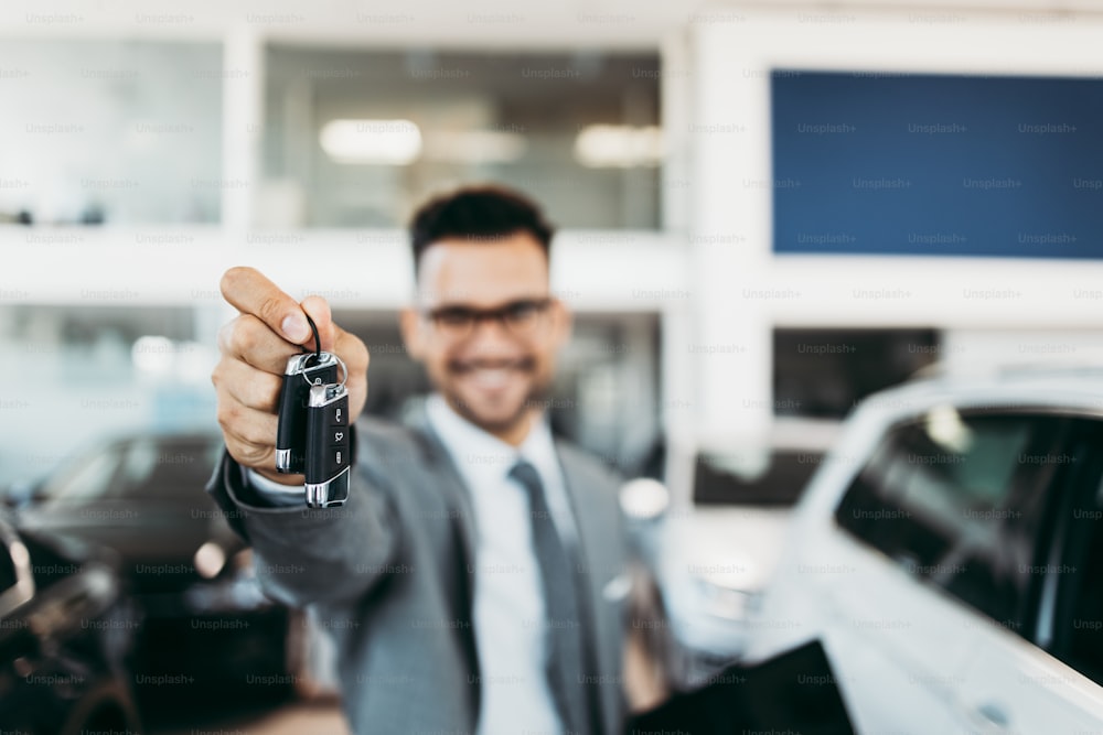 Good looking, cheerful and friendly salesman holding keys and posing in a car salon or showroom.