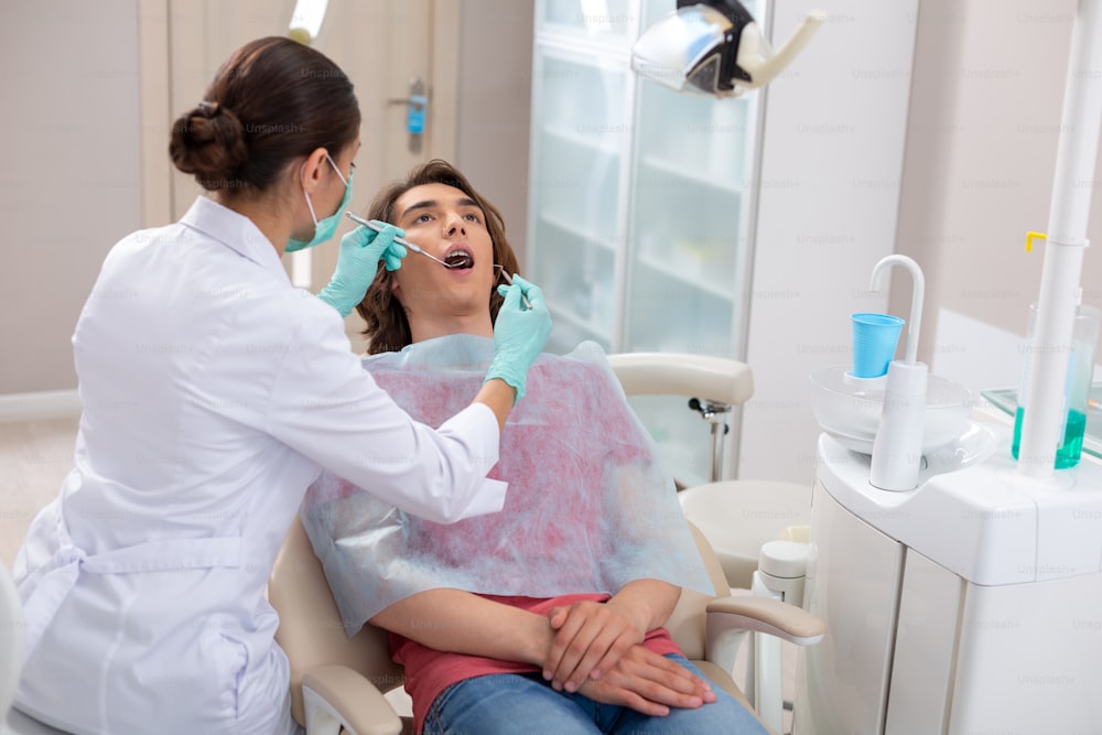 중괄호를 가진 십대. 치과 진료소에서 치과 교정기로 남성 환자의 치아를 검사하는 여성 치과 의사