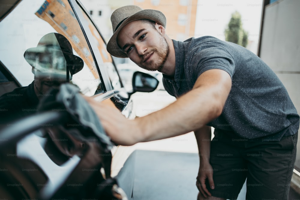 Giovane bell'uomo con cappello che pulisce l'auto con lo straccio, concetto di dettaglio dell'auto (o valeting).