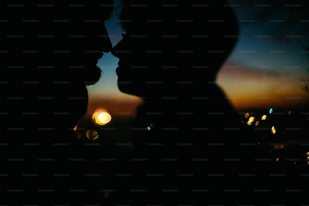 夕焼けの空と街の明かりを背景に、お互いを見つめ合い、優しく触れ合う幸せなカップルのシルエット