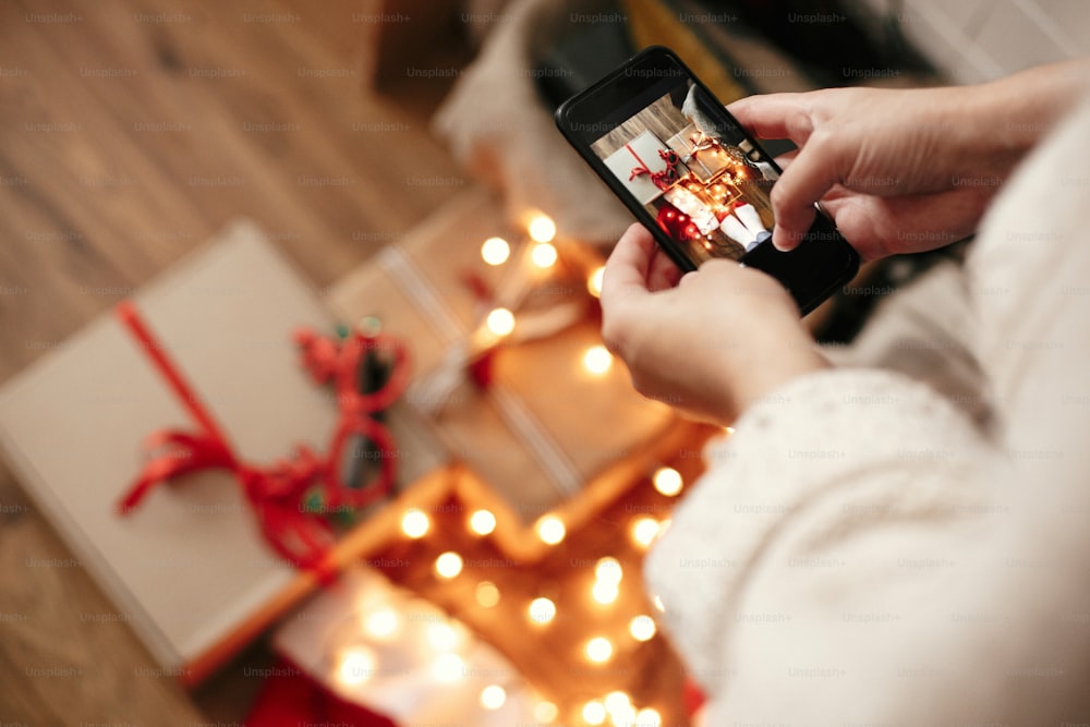 Mãos segurando telefone e tirando foto de caixas de presente de Natal, chapéu de Papai Noel, luzes de iluminação no fundo de madeira no quarto escuro. Menina hipster elegante no suéter que faz a foto do leigo plano do Natal