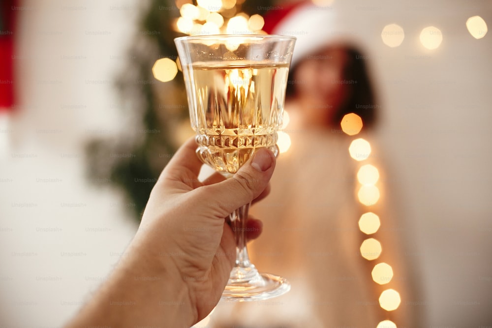 Man toasting with champagne glass to happy girl in santa hat with sparkler, celebrating at christmas tree lights in festive room. Happy New Year eve party. Happy Holidays
