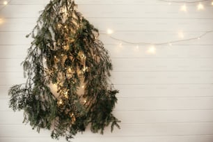 Stylish modern christmas tree made of fir branches, hanging on white wall with festive garland light. Eco Zero waste decoration of a room for christmas holidays. Merry Christmas. Happy Holidays