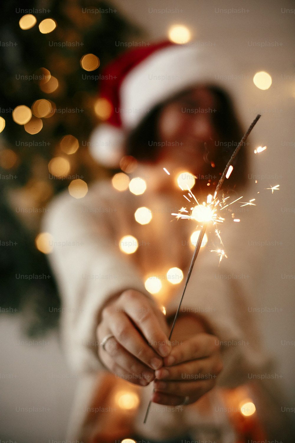 暗い部屋のモダンなクリスマスツリーの光を背景に、サンタの帽子をかぶったスタイリッシュな女の子の手で燃えている大きな線香花火。花火を持つ女性。大晦日あけましておめでとうございます。メリークリスマス