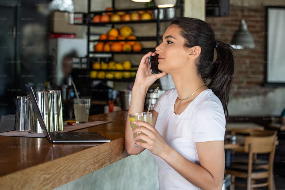 Mujer joven encantadora que llama con teléfono celular mientras está sentada sola en la cafetería durante el tiempo libre, mujer atractiva con una sonrisa linda que tiene una conversación que habla con el teléfono móvil mientras descansa en el bar