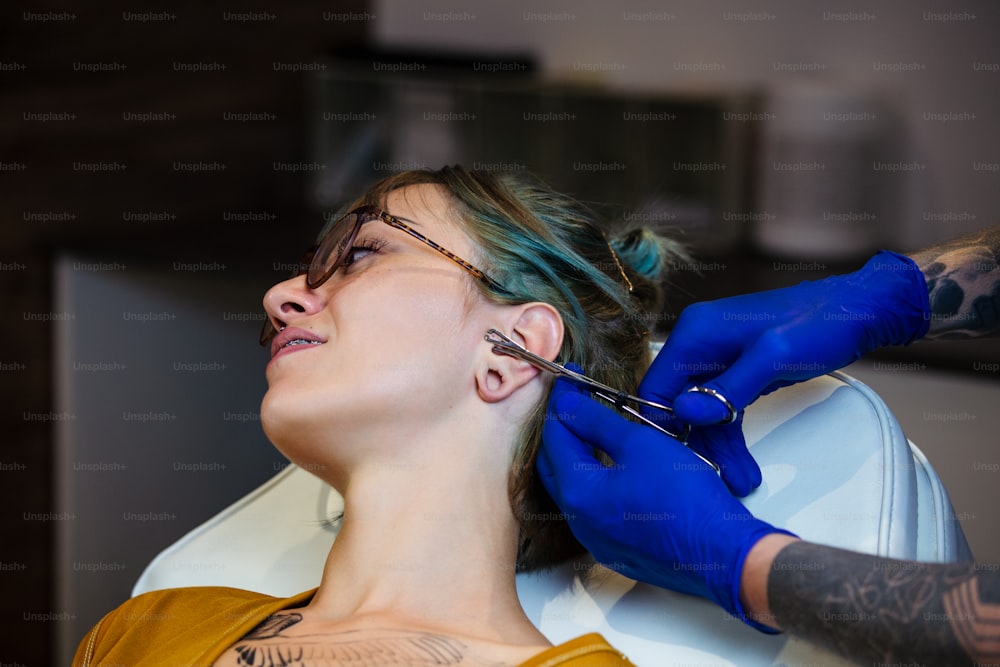 Jeune femme se faisant percer l’oreille. Homme montrant un processus de perçage avec du matériel médical stérile et des gants en latex. Procédure de perçage corporel
