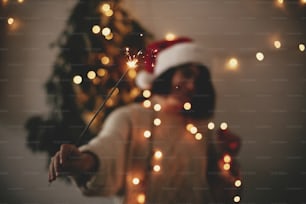 Grande scintilla che brucia in mano di elegante ragazza hipster in cappello da babbo natale sullo sfondo della luce moderna dell'albero di Natale nella stanza buia. Atmosfera di Felice Anno Nuovo. Buon Natale. Buone Feste