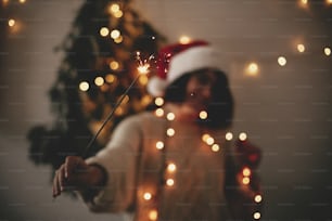 Big sparkler burning in hand of stylish hipster girl in santa hat on background of modern christmas tree light in dark room. Happy New Year eve atmosphere. Merry Christmas. Happy Holidays