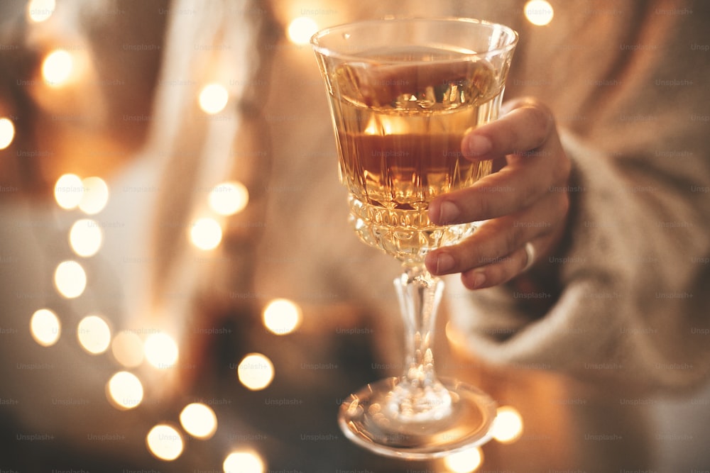 Champagne glass in hand of stylish girl in cozy sweater with christmas lights, celebrating and toasting in dark festive room. Happy New Year eve party. Happy Holidays. Merry Christmas