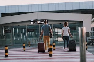 Retrato de cuerpo entero de la vista trasera del caballero y la dama que llevan sus maletas con ruedas y se dirigen a la entrada de la terminal del aeropuerto