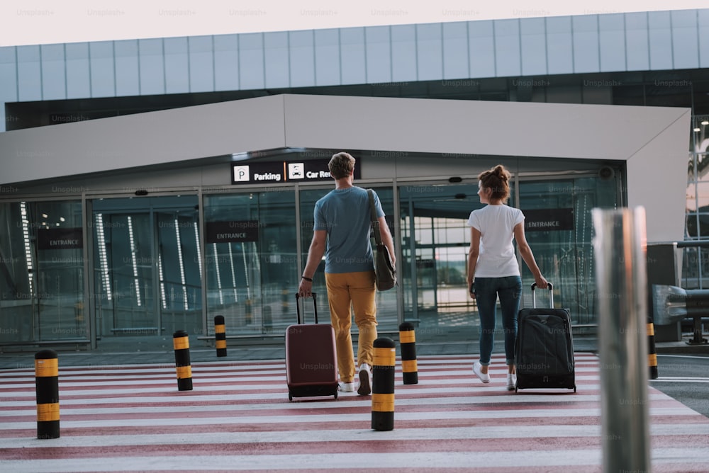 Rückansichtsporträt in voller Länge von Herrn und Dame, die ihre Trolley-Taschen tragen und zum Eingang des Flughafenterminals gehen
