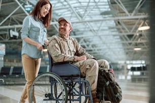 Low angle of disabled Caucasian soldier sitting in wheelchair. Young couple is smiling while talking to each other. Homecoming and military concept