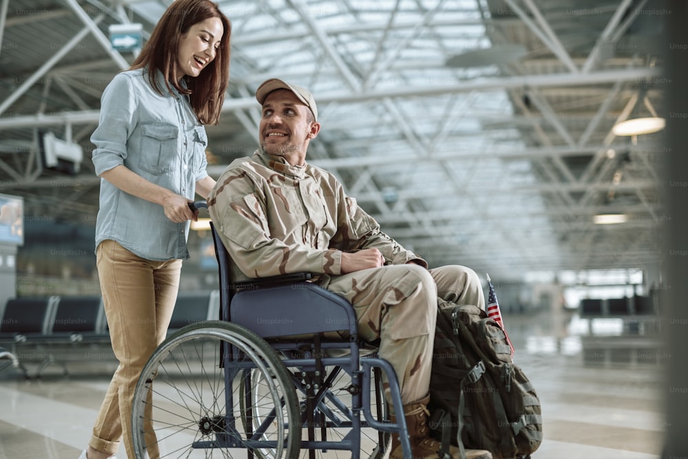 Low angle of disabled Caucasian soldier sitting in wheelchair. Young couple is smiling while talking to each other. Homecoming and military concept