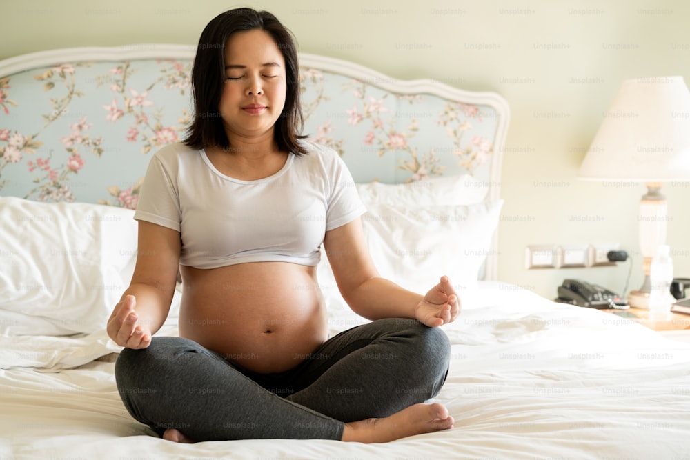 Mulher grávida fazendo exercício de ioga na cama no quarto em casa enquanto cuida de seu filho. A jovem mãe feliz esperando segurando o bebê na barriga grávida. Maternidade pré-natal e gravidez.