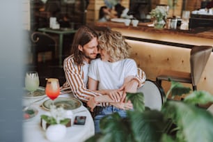 Bearded young man embracing his charming girlfriend from behind while she smiling. They sitting at the table with plates and cocktails