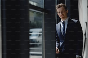 Taille hoch, attraktiver eleganter Geschäftsmann im dunklen Anzug, der in der Nähe des Fensters posiert und lächelt