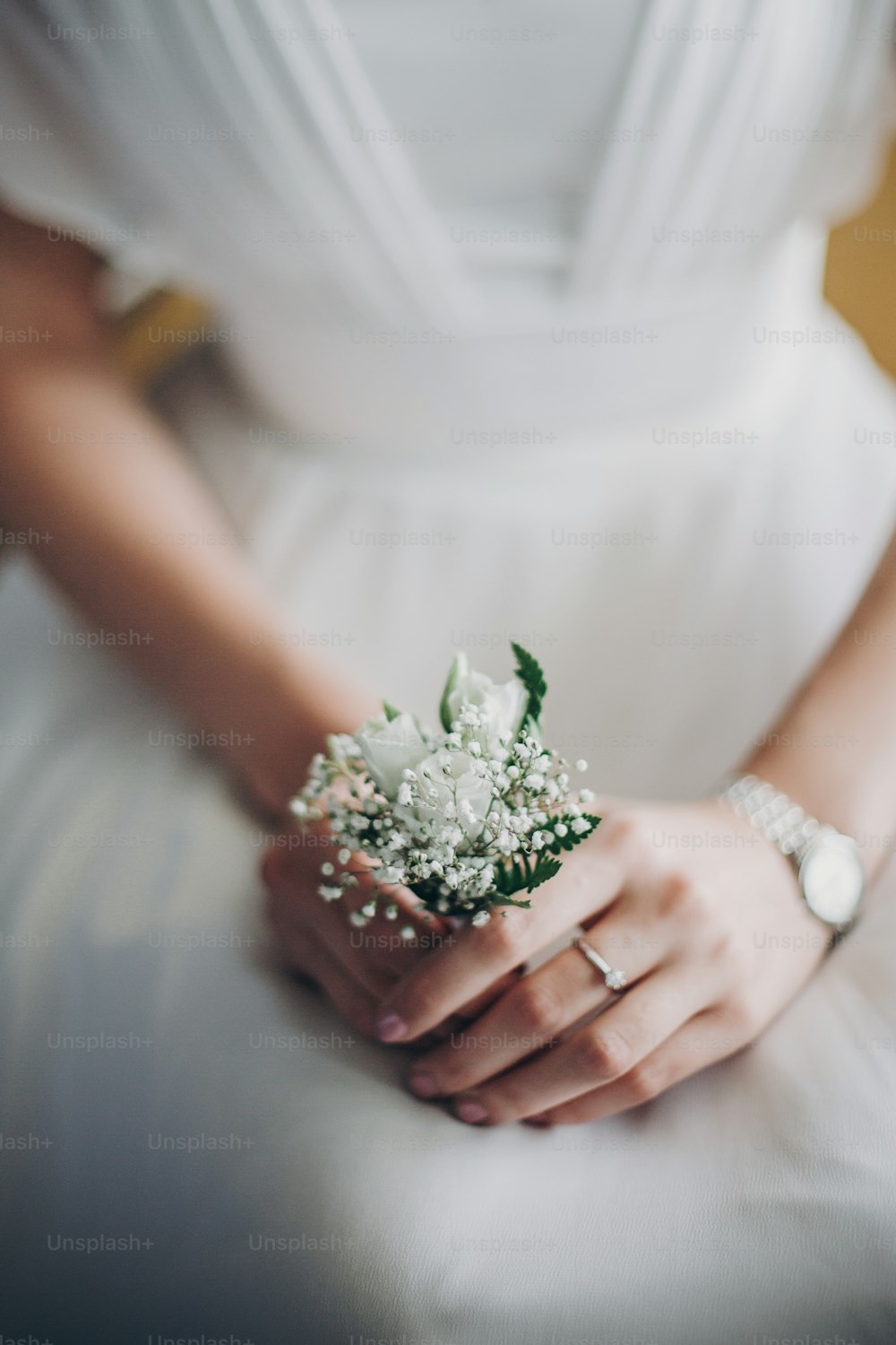 Stylish bride holding in hands boutonniere with white roses and gypsophila for groom in morning light in hotel room. Morning preparations