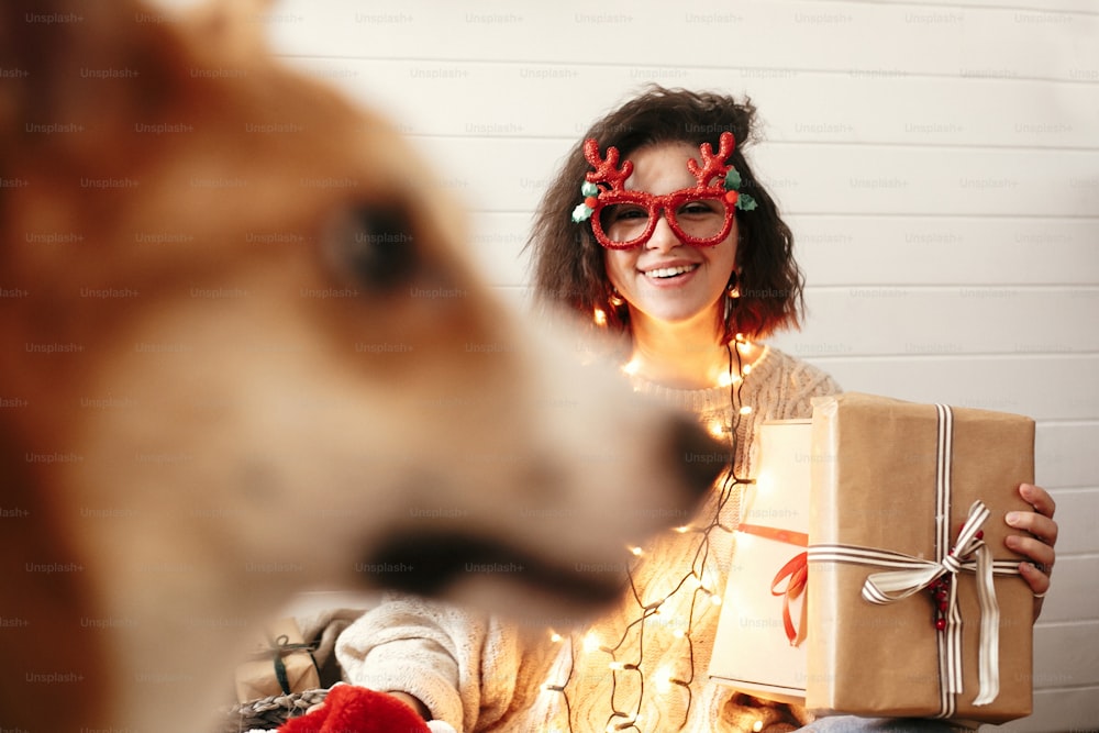 크리스마스 조명에 크리스마스 선물 상자를 들고 귀여운 황금 개에게 웃고 있는 세련된 행복한 소녀. 사슴 뿔과 아늑한 스웨터를 입고 크리스마스 선물을 보여주는 축제 안경을 쓴 젊은 여자