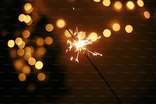 Hand holding glowing sparkler on background of golden christmas lights, celebrating in dark festive room. Space for text. Happy New Year eve party. Happy Holidays. Fireworks burning in hand