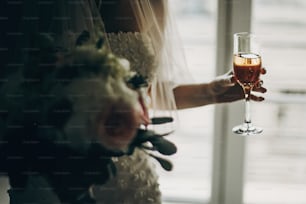 Zugeschnittene Ansicht der stilvollen Braut, die einen modernen Hochzeitsstrauß hält und mit Champagnerglas in der Nähe des Fensters im Hotelzimmer anstößt. Wunderschöne Brautmorgenvorbereitung vor der Hochzeitszeremonie