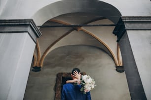 Stylish bride holding bouquet and gently hugging groom neck in city street, back view. Gorgeous wedding couple of newlyweds embracing in old buildings. Romantic moment