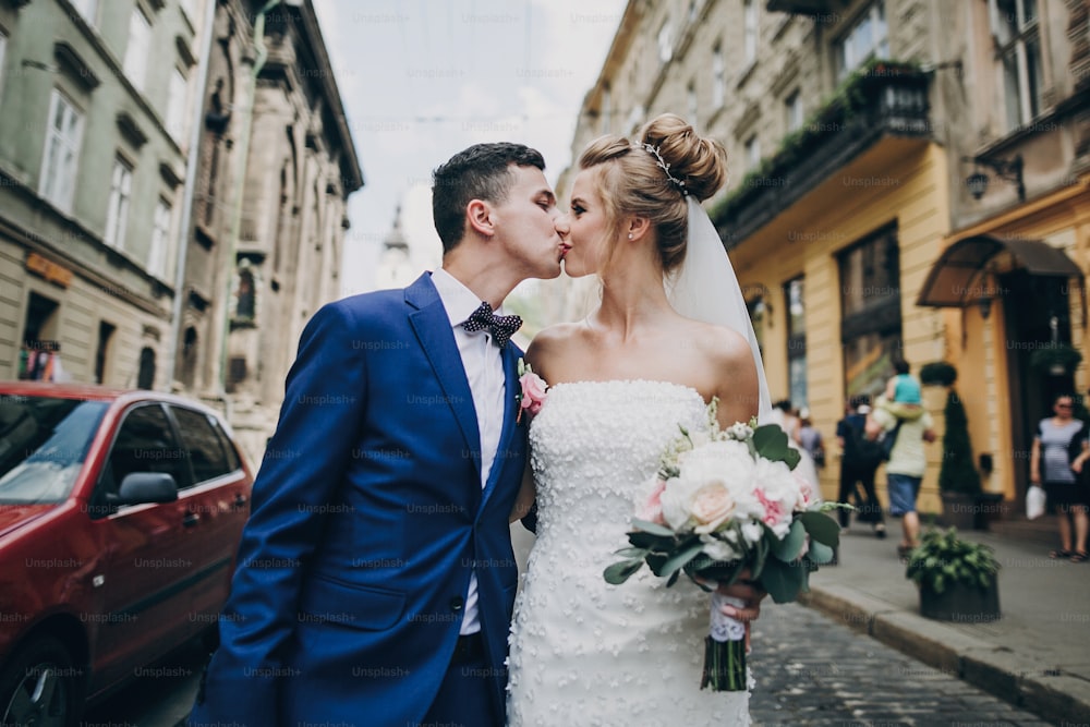 Stylish happy bride and groom walking and kissing in sunny city street. Gorgeous wedding couple of newlyweds embracing outdoors. Romantic moment
