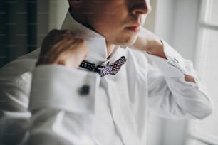 Stylish groom in white shirt putting on bow tie near window in hotel room. Morning preparation before wedding ceremony. Man getting ready before luxury event