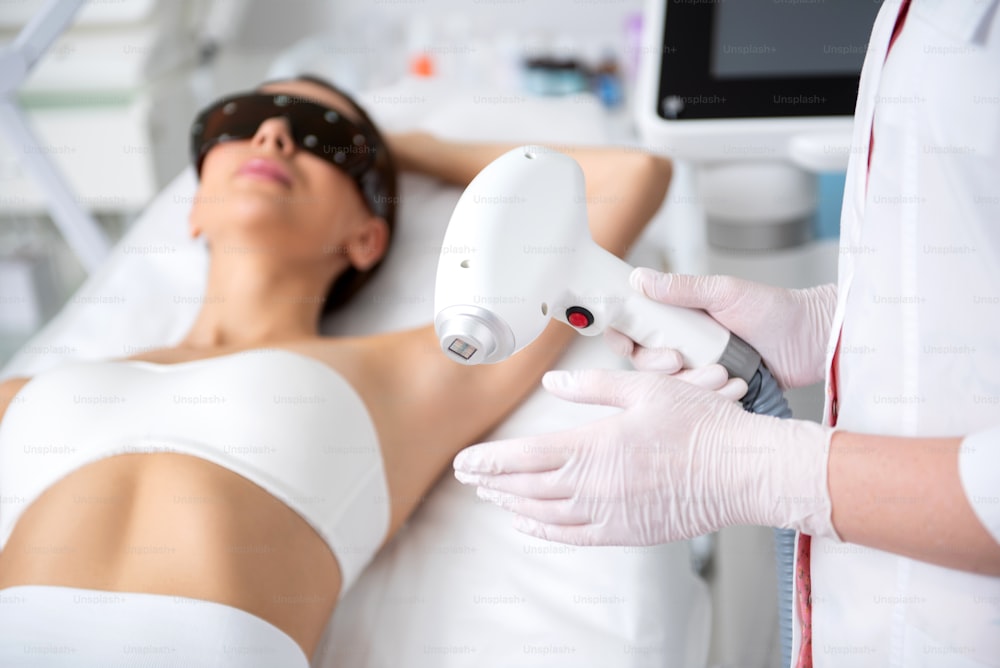 Aesthetic body treatment. Close up side on portrait of cosmetologist holding laser hair removal device in hand while young woman in eyeglasses lying on couch