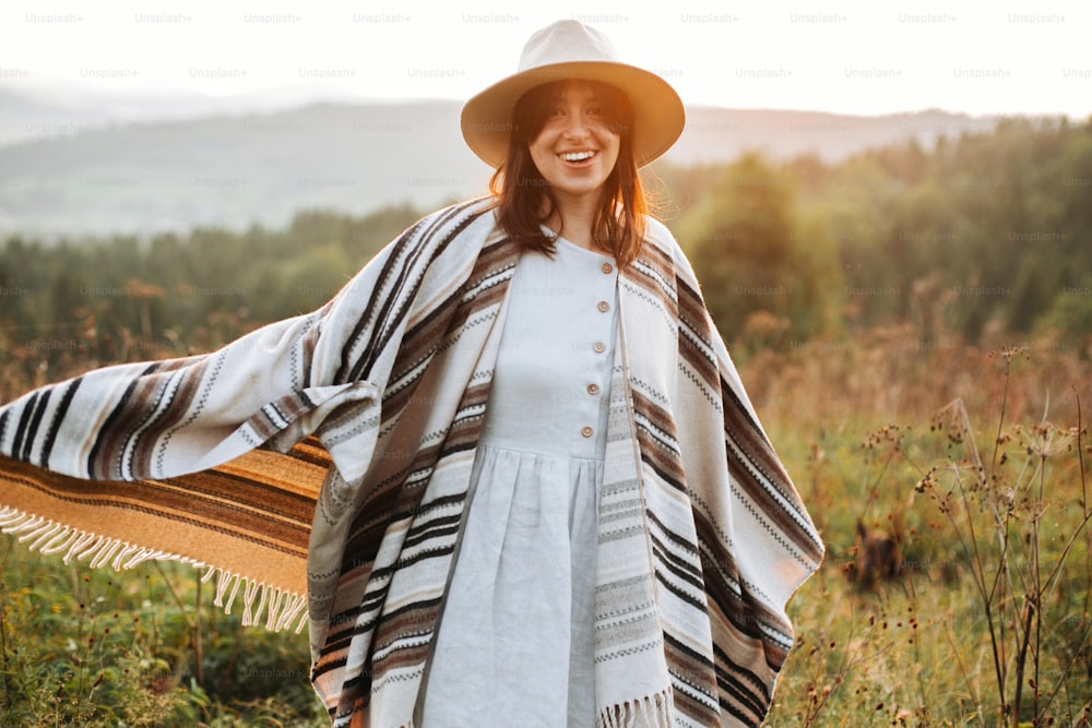 Stylish boho girl smiling  in sunny light at atmospheric sunset in meadow. Happy hipster woman in poncho and hat enjoying traveling in evening mountains. Space for text