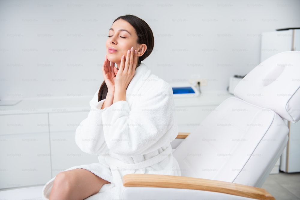 Health and body treatment. Waist up side on portrait of pretty smiling brunette woman sitting on couch in spa center and touching her face satisfied closing eyes