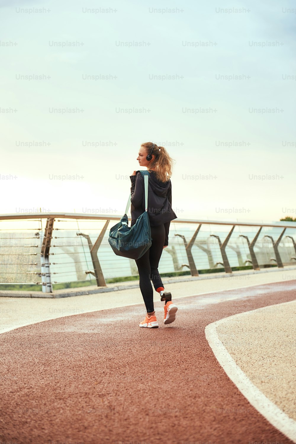Morning workout. Back view of young disabled woman in sports clothing with leg prosthesis holding her sport bag while standing on the bridge in the morning. Disabled sport concept. Motivation. Healthy lifestyle