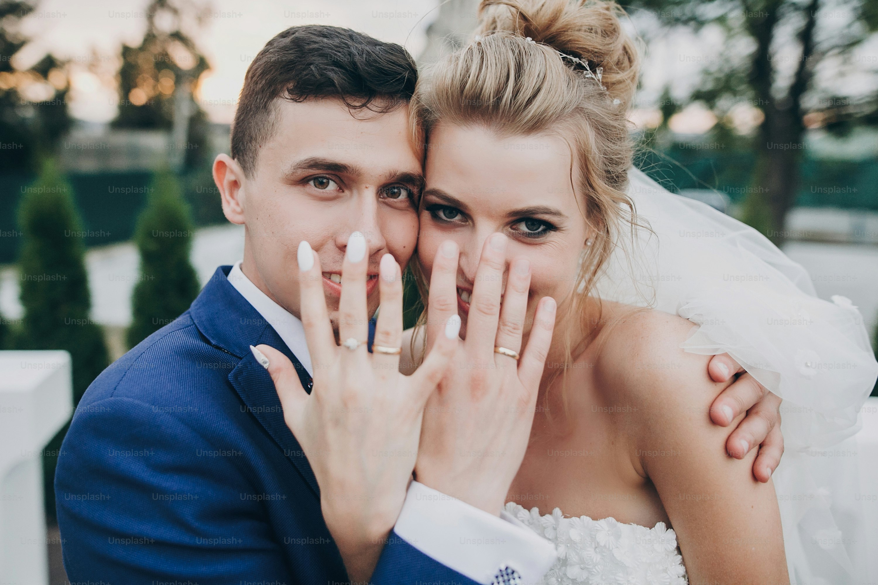 Just married. Stylish happy bride and groom showing hands with wedding rings at wedding reception outdoors. Gorgeous wedding couple of newlyweds having fun