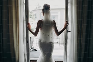 Silueta de la elegante novia que abre el balcón de la ventana con luz suave en la habitación del hotel. Espalda de hermosa novia sensual en vestido blanco. Preparación matutina antes de la ceremonia de la boda