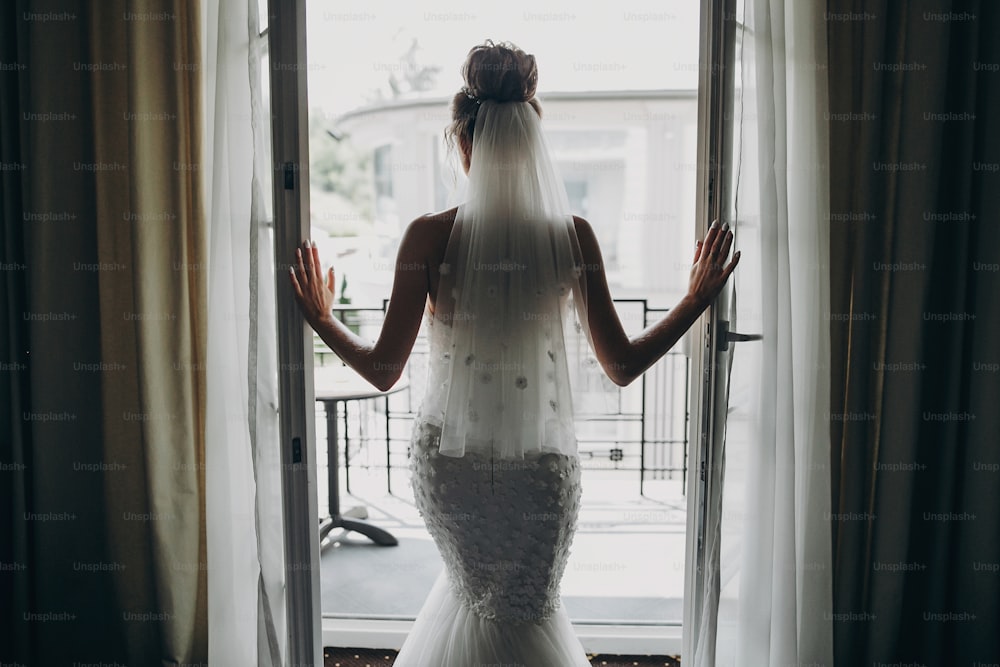 Silhouette des stilvollen Brautfensters Balkon in weichem Licht im Hotelzimmer. Rückseite der wunderschönen sinnlichen Braut in weißem Kleid. Morgenvorbereitung vor der Hochzeitszeremonie