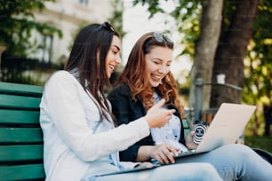 Two beautiful young girlfriends laughing while looking at a laptop screen sitting on a bench in the park drinking coffee .