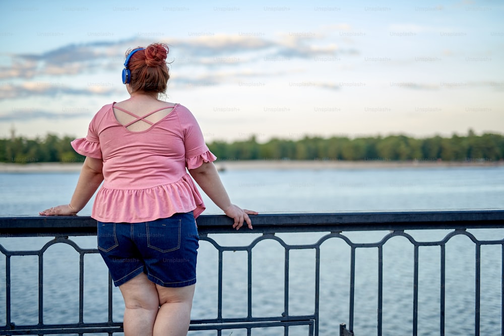 Young and fat woman looking at river in headphone and listening music