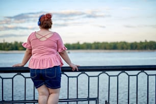 Young and fat woman looking at river in headphone and listening music