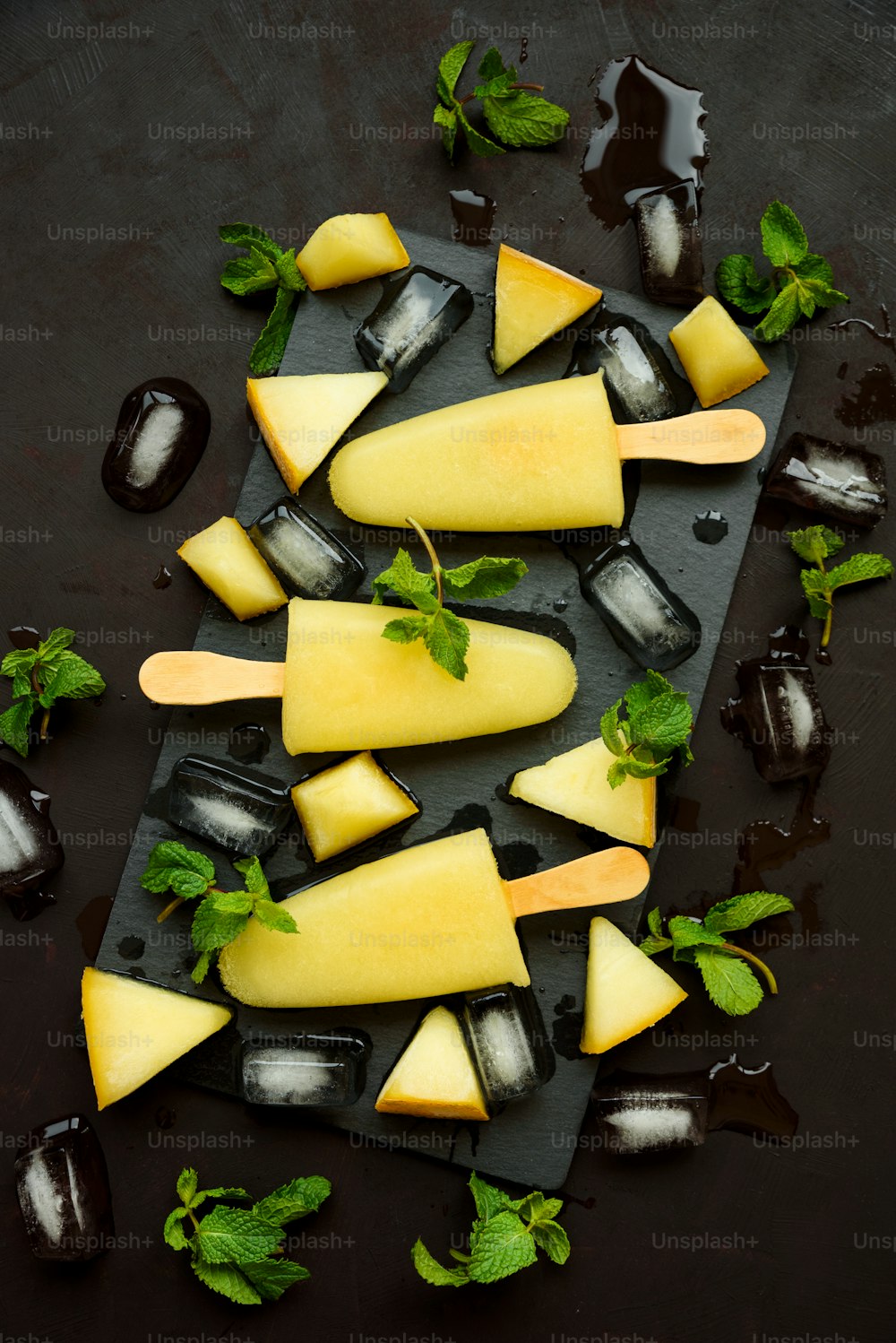 Homemade melon ice cream, pops or popsicles with ice, melon slices and mint on rectangular dark slate plate. Summer food. Flat lay. Top view