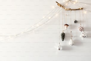 Juguetes vintage navideños que cuelgan de una rama de madera en una pared blanca con luces festivas en una habitación moderna, estilo minimalista escandinavo. Elegantes adornos de vidrio, decoraciones navideñas. Espacio para el texto
