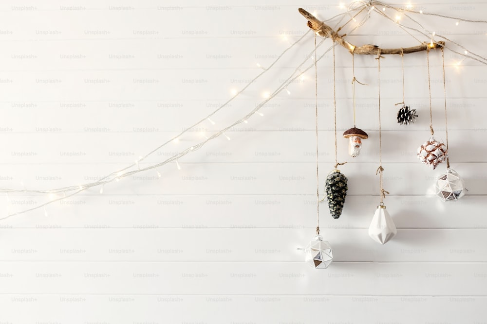 Weihnachts-Vintage-Spielzeug hängt an Holzzweig an weißer Wand mit festlichen Lichtern in modernem Raum, skandinavischer Minimal-Stil. Stilvolle Glasornamente, Urlaubsdekorationen. Platz für Text