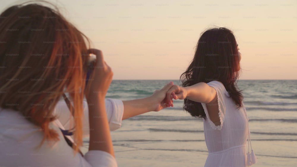 Young Asian lesbian couple using camera taking photo each other near beach. Beautiful women lgbt couple happy romantic moment when sunset in evening. Lifestyle lesbian couple travel on beach concept.