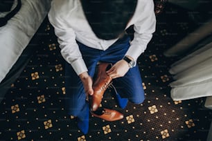 ホテルの部屋の窓の近くで茶色の靴を履く青いスーツを着たスタイリッシュな新郎、上面図。結婚式前の朝の準備。贅沢なイベントの前に準備する男